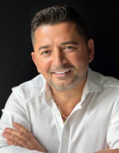 Smiling man with short dark hair and light stubble, wearing a white shirt, arms crossed against a dark background.