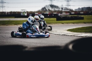 abcdhe | Tony J. Selimi Three go-karts and drivers in helmets navigate a left-hand turn on an outdoor track, competing closely for position.