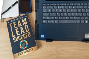 Desk top view with a laptop keyboard on the right, a leadership book titled 'Team Lead Succeed' by Nick Fewings on the left, a pen, notebook, and a blue-cased smartphone on a wooden surface.