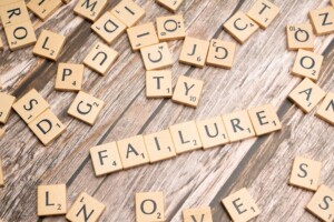 Scrabble tiles spell 'FAILURE' on a wooden tabletop with scattered letters around.