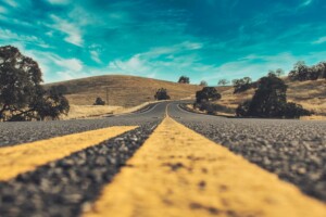 image-15 | Tony J. Selimi Two yellow center lines on a winding rural highway disappearing toward rolling hills under a blue sky.