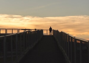 Silhouette of a person standing at the end of a walkway with railings on both sides during sunset at golden hour.